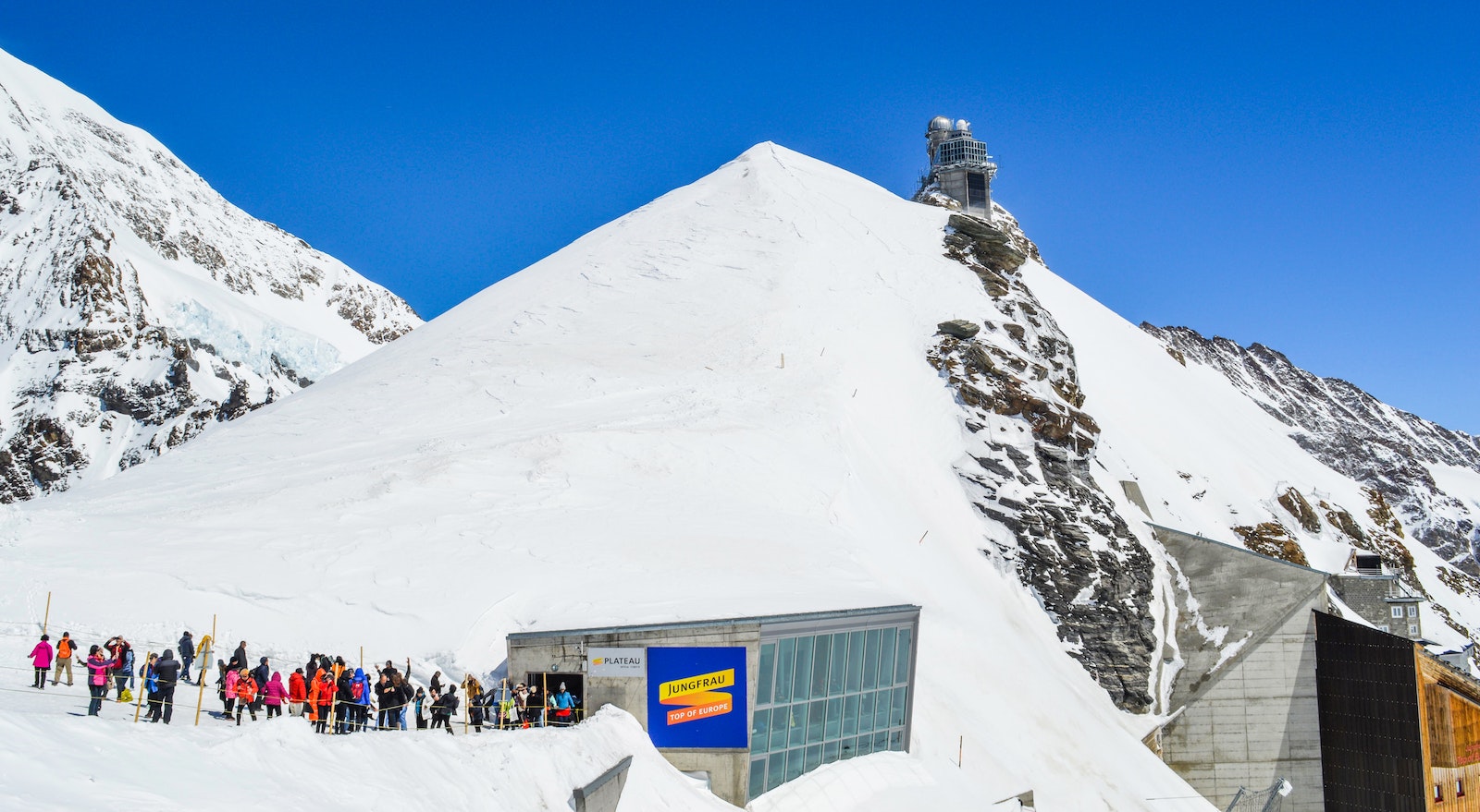 Anonymous group of tourists with skis near old building on snowy mountain under blue sky