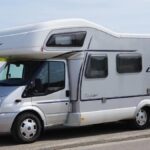 Camping-car de classe moyenne stationné au bord d'une route, paysage agricole avec champ de colza en arrière-plan