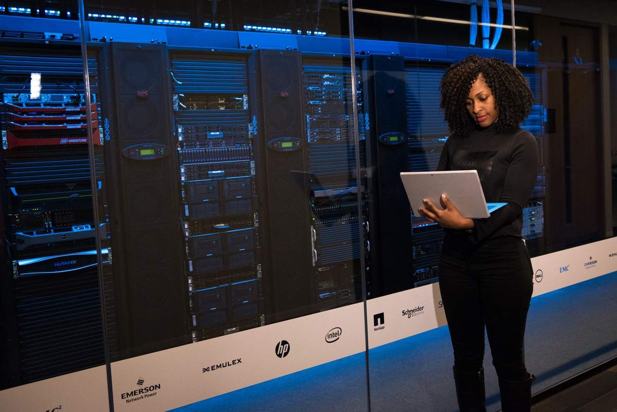 Femme consultant une tablette devant une baie de serveurs informatiques illuminée en bleu Femme consultant une tablette devant une baie de serveurs informatiques illuminée en bleu