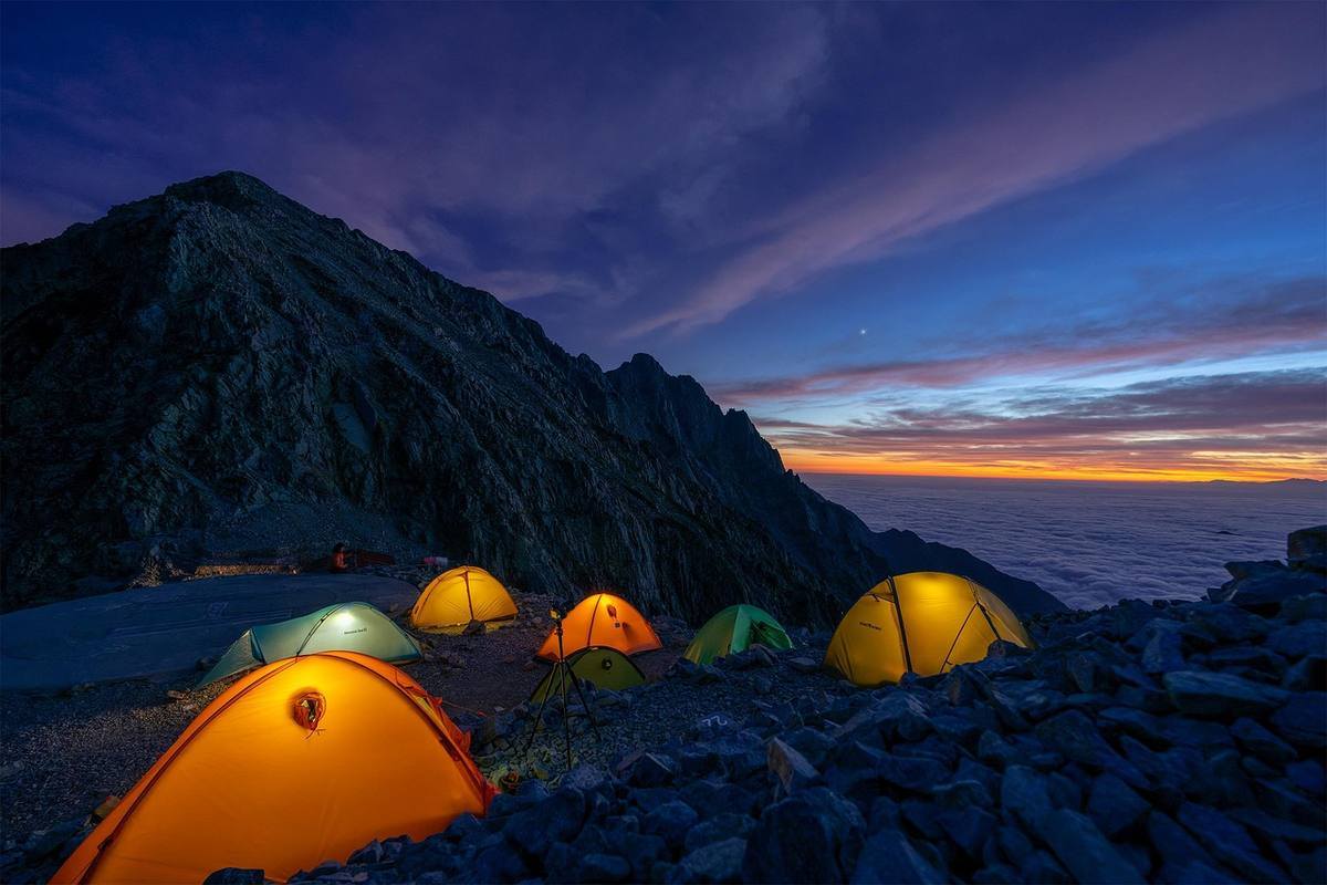 Plusieurs tentes colorées illuminées de l'intérieur, campement nocturne en montagne avec mer en arrière-plan