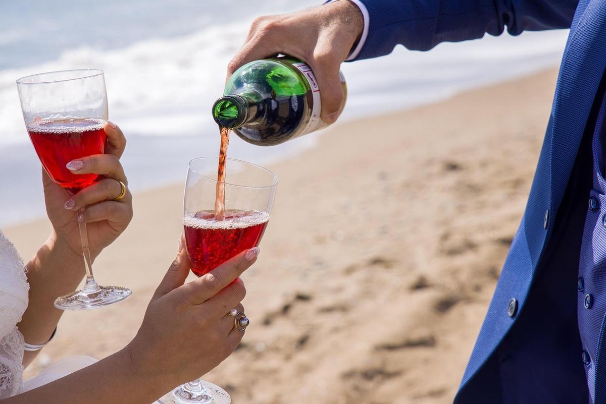 Une personne verse un liquide rouge dans un verre, une autre tient un verre rempli, sur une plage avec l'océan en arrière-plan