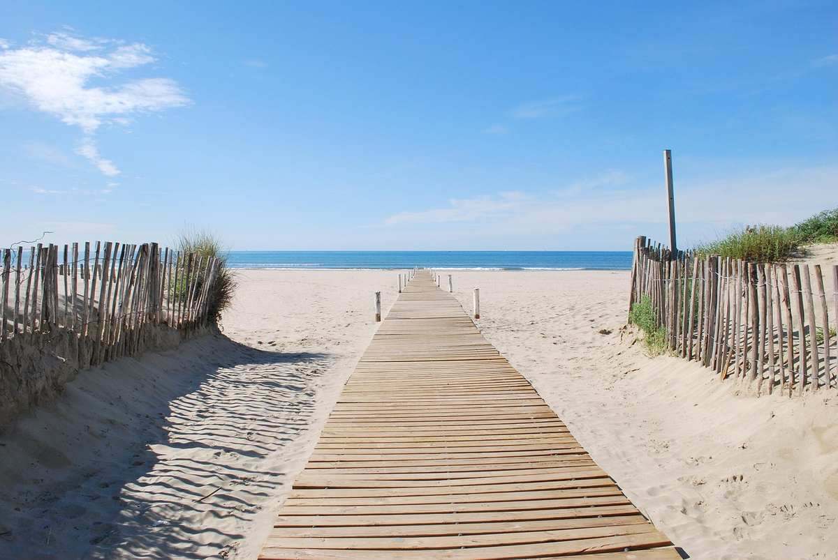 Passerelle en bois sur la plage, bordée de clôtures, menant à la mer sous un ciel bleu Passerelle en bois sur la plage, bordée de clôtures, menant à la mer sous un ciel bleu